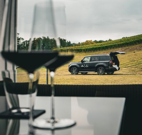 A pair of wine glasses in soft focus in the foreground, framing a black Lexus SUV parked on a grassy hillside vineyard in the distance, with rolling rows of vines beneath an overcast sky.