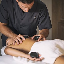 Massage therapist performing a hot stone massage on a client's back in a spa setting.