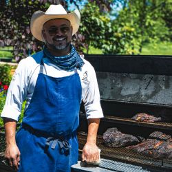 Chef wearing a cowboy hat and blue apron stands beside an outdoor smoker grill with slow-cooked meats, surrounded by lush greenery on a sunny day.