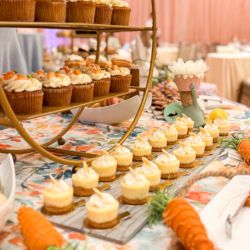 Assorted spring desserts arranged on a decorative table display, featuring carrot cake cupcakes and miniature cheesecakes with piped frosting, surrounded by fresh carrots and pastel décor.